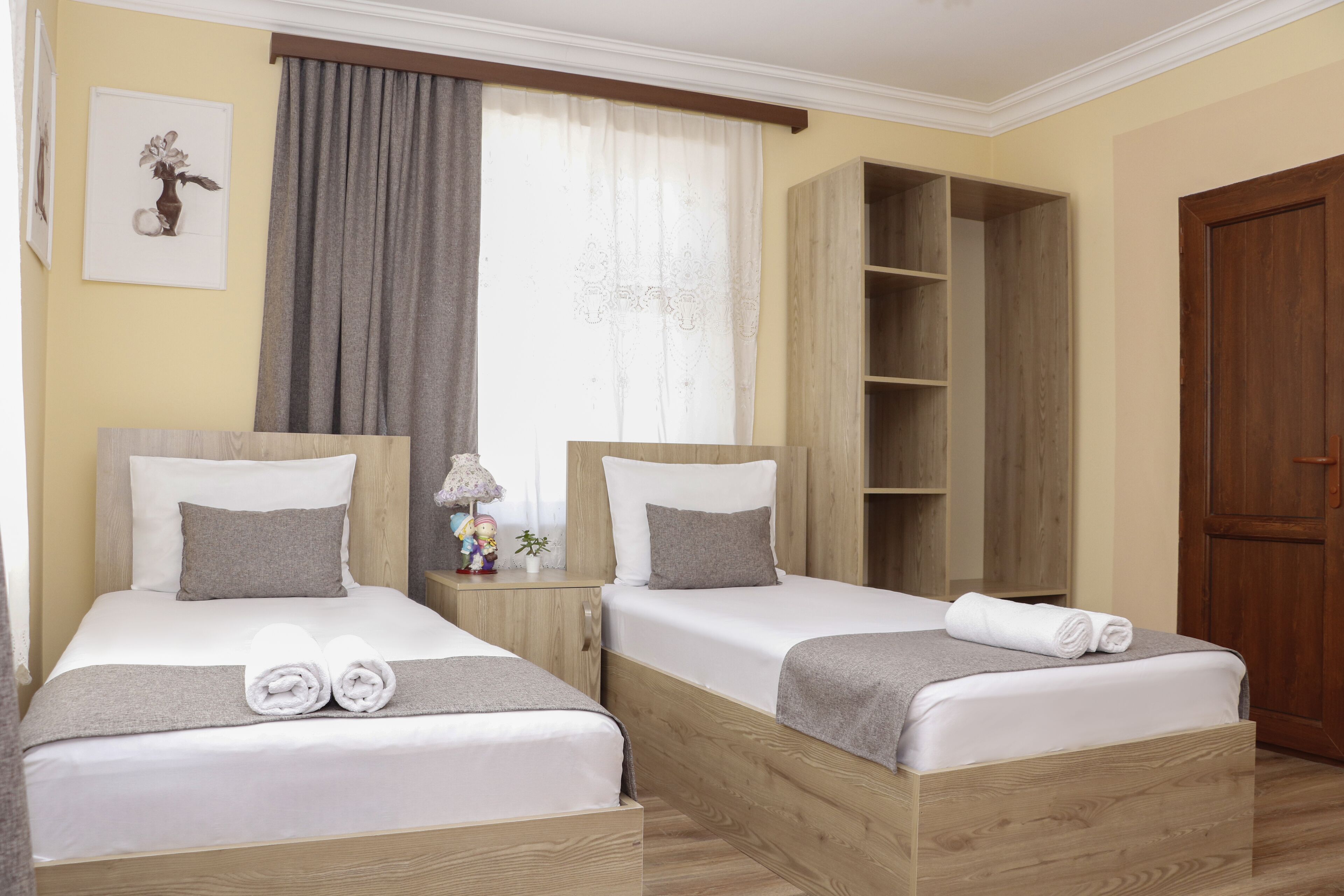 A hotel room with two beds, a wooden wardrobe, a small table with a lamp, and a large window with curtains.