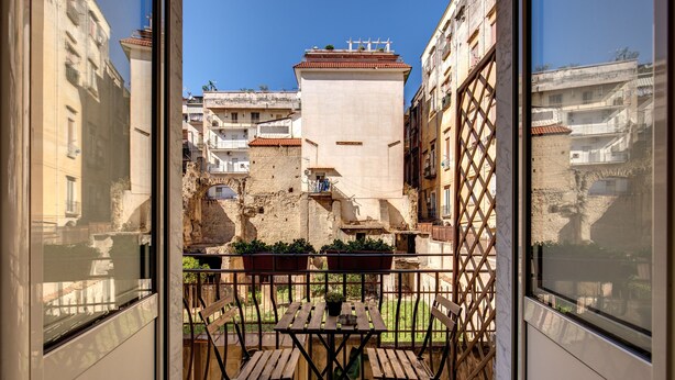 Un balcón con mesa y sillas de madera, plantas en macetas y vistas a edificios históricos y un puente de piedra.