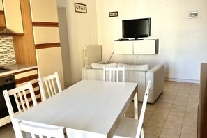 A compact living space with a dining table, white chairs, a sofa, and a television mounted on the wall.
