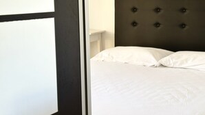 Un llit amb capçalera amb borlonis, roba de llit blanca i una vista parcial d'una paret blanca i un marc de porta fosc.