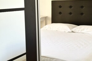Un lit doté d’une tête de lit capitonnée, de draps blancs et offrant une vue partielle sur un mur blanc et le cadre d’une porte de couleur foncée.