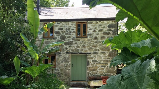 Een stenen huis met een groene deur, omringd door weelderig groen en een stenen pad dat naar de ingang leidt.