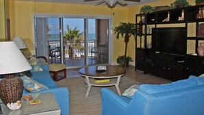 A living room with a sofa, a coffee table, a TV, and a bookshelf. There is a balcony with a view of the ocean. - Enjoy your beach holiday in this updated oceanfront two bedroom, two bath condo. (New Smyrna Beach)