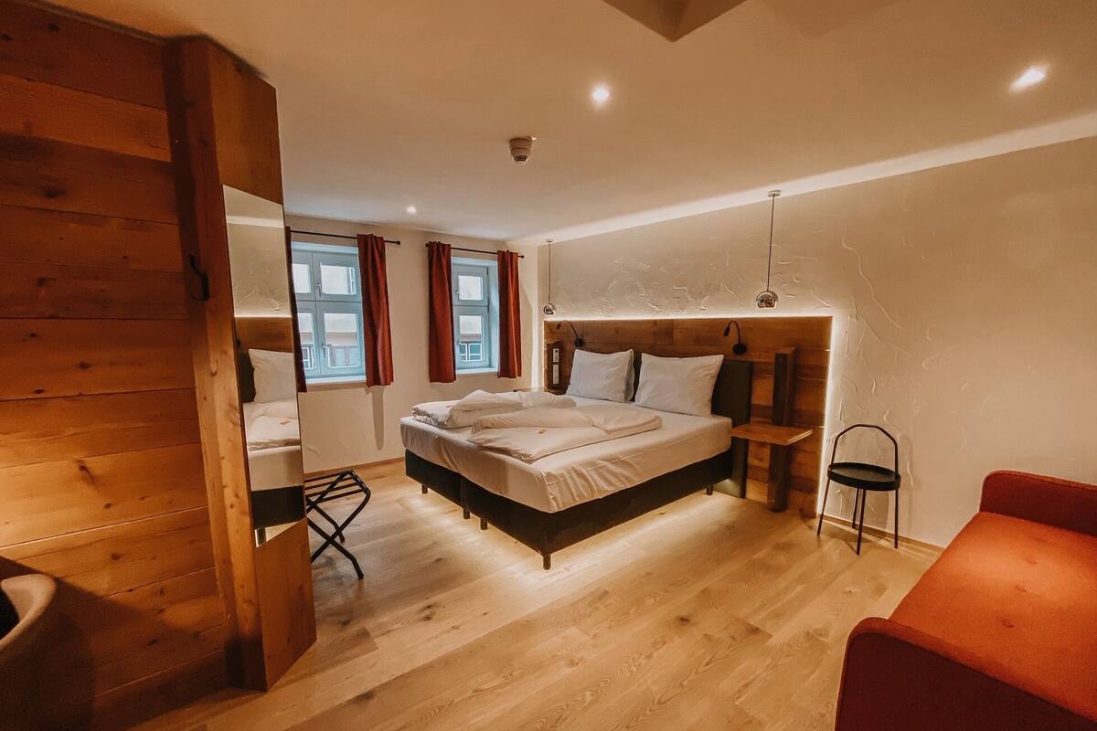 A bedroom with a wooden floor, a bed with white bedding, a red chair, and a small table.