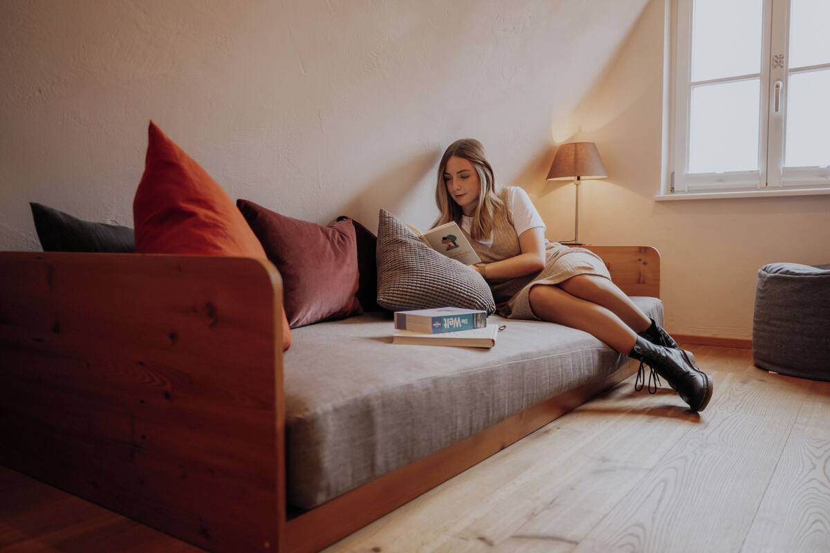 A person sitting on a sofa with a book, a lamp, and a window in the background.