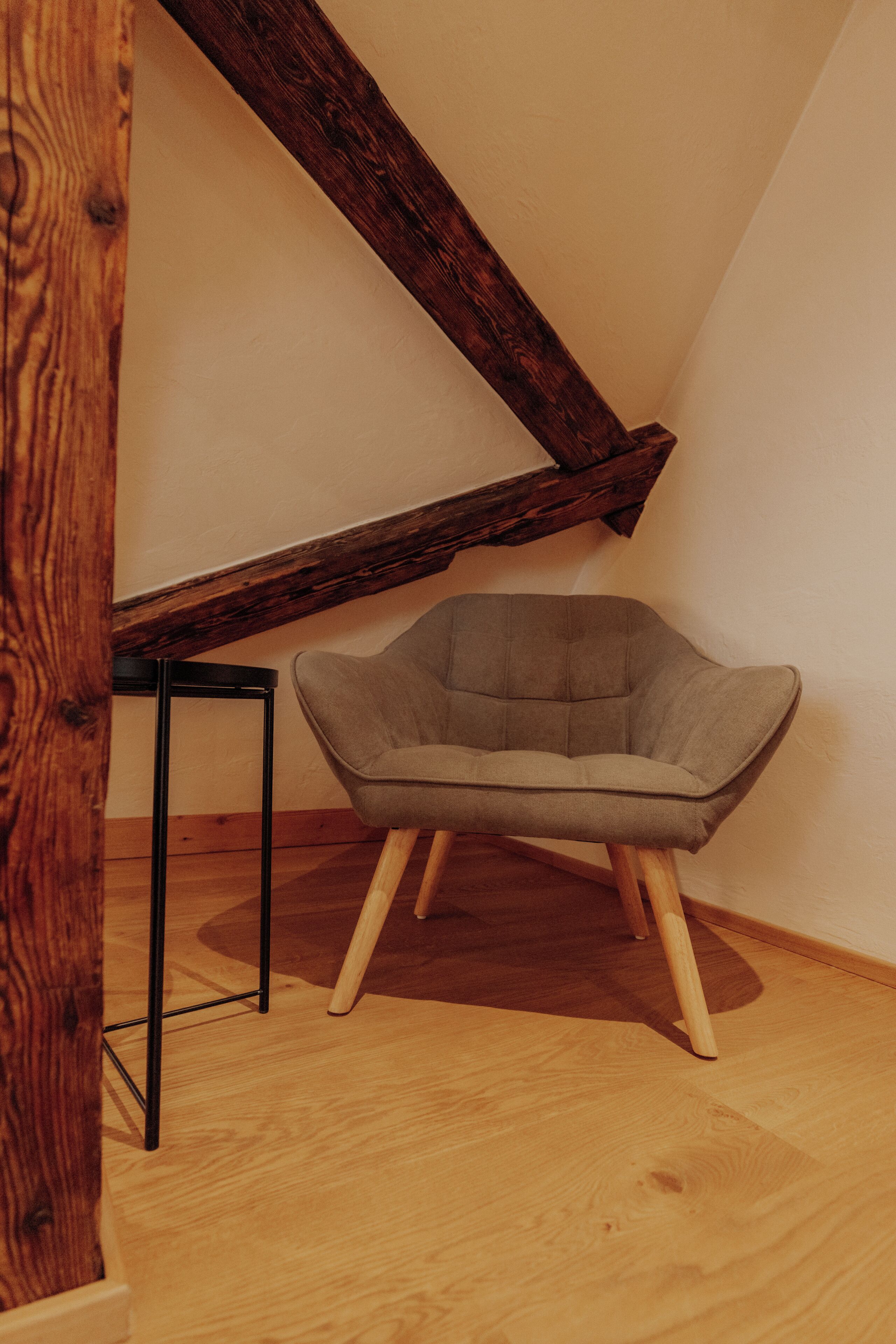 A corner room with a wooden floor, a grey upholstered chair, and a black metal table.