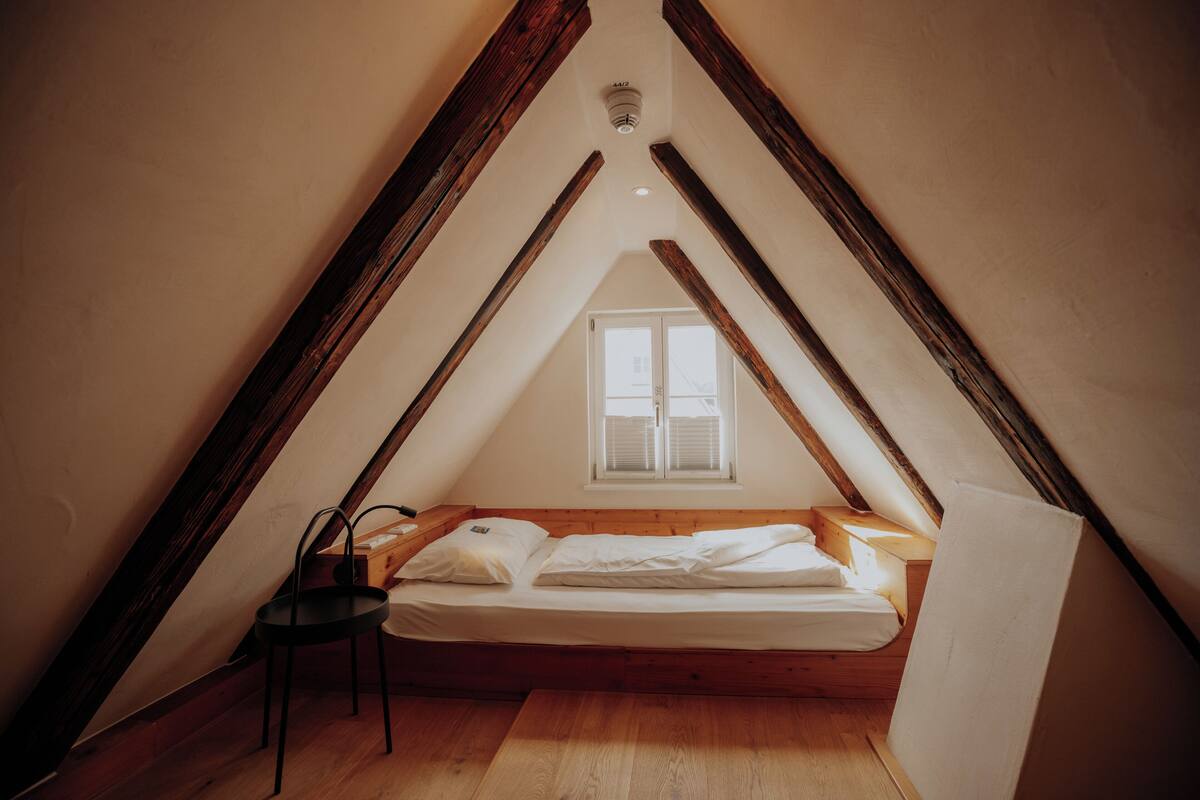 A small, triangular attic bedroom with a single bed, a window, and wooden beams.