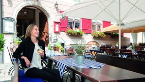 Un café al aire libre con mesas y sillas, una mujer sentada en una mesa con una bebida, y un edificio con contraventanas rojas al fondo.