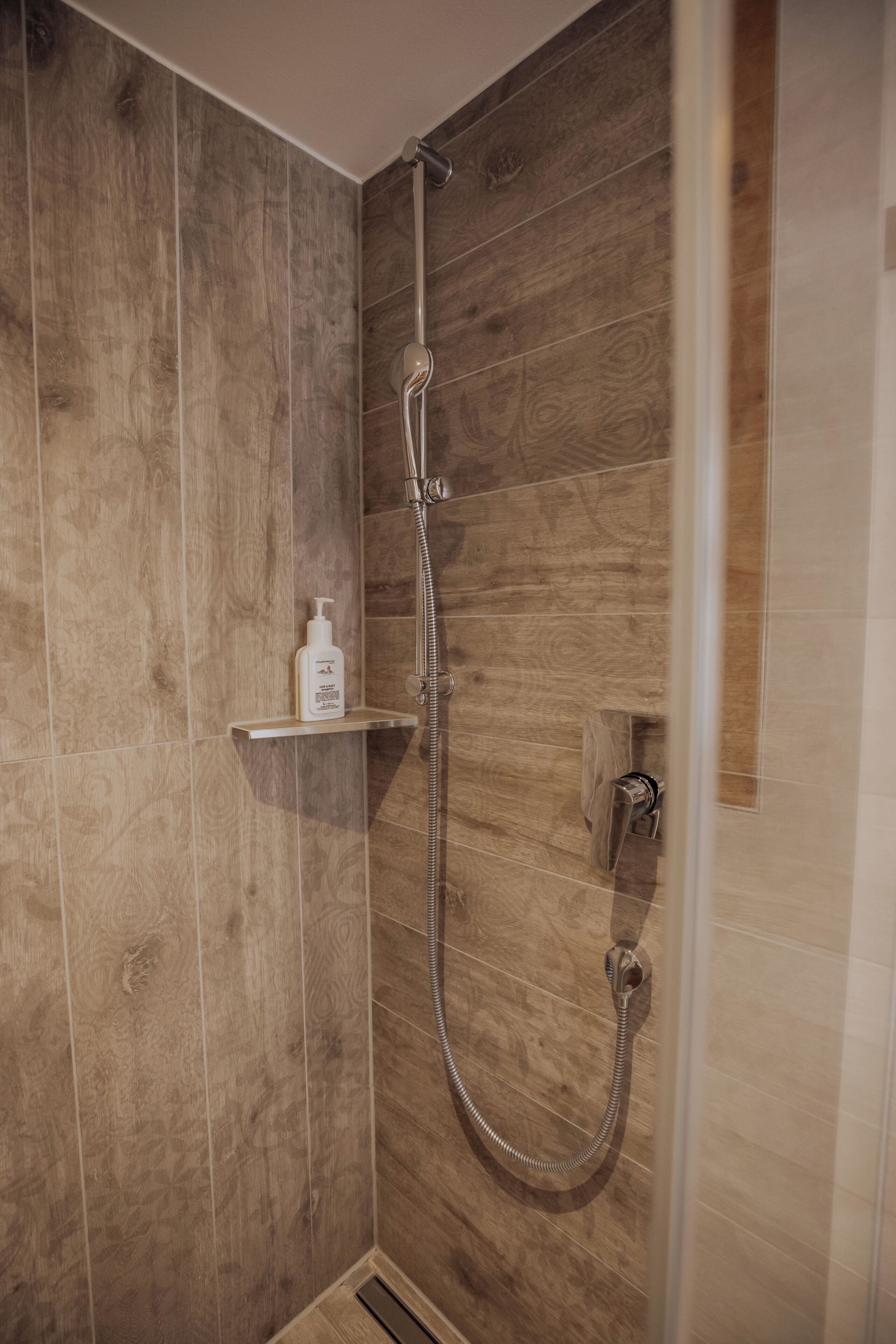 Ducha con pared de madera, cabezal de ducha manual y una botella de champú en un estante.