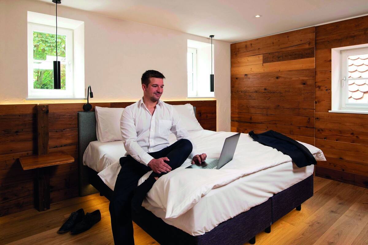 A man sitting on a bed with a laptop, a wooden wall, and a window with a view of greenery.