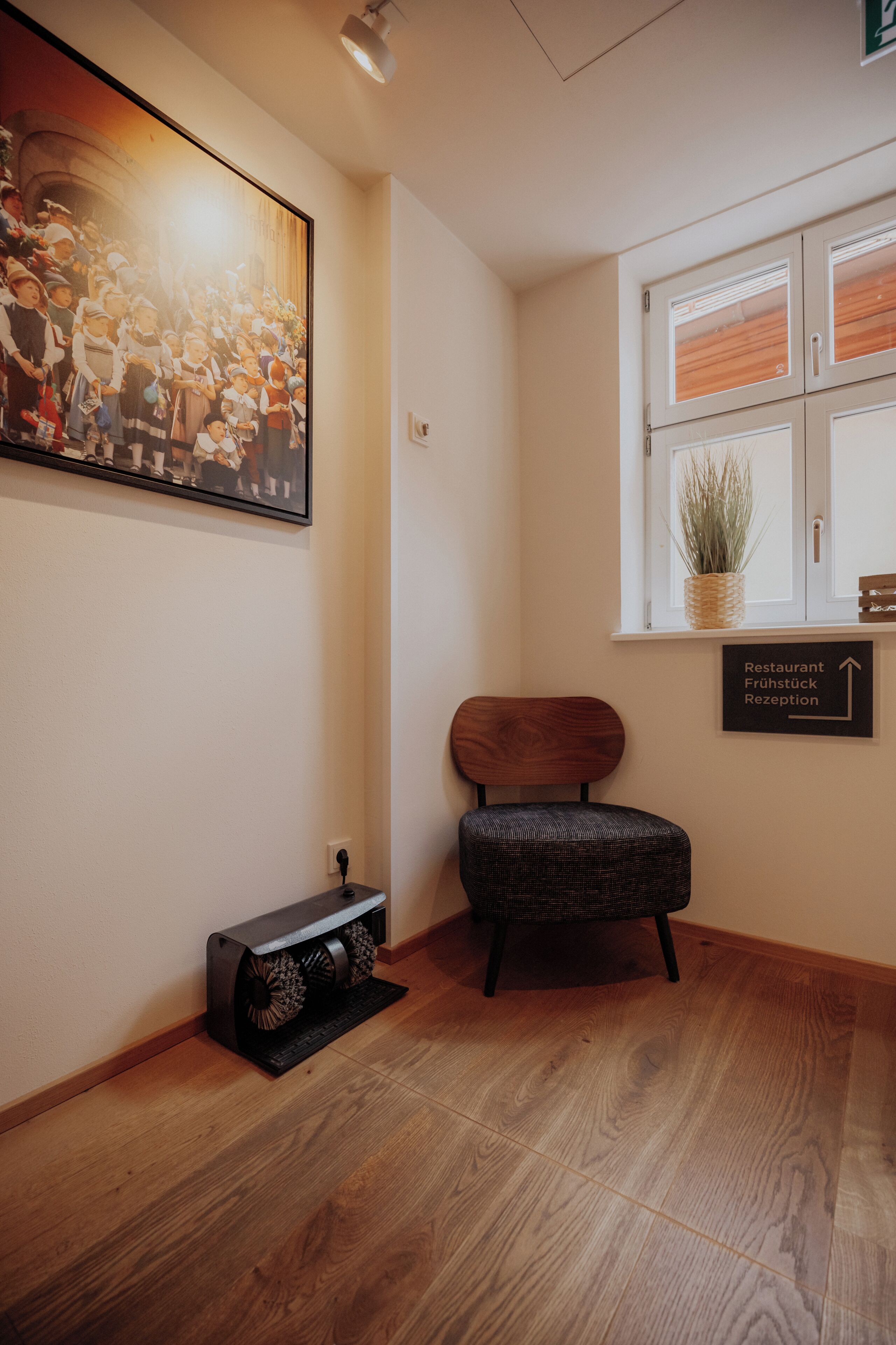Un área de recepción con suelo de madera, una fotografía enmarcada en la pared, una planta en maceta y un letrero que indica el camino al restaurante y a la recepción.