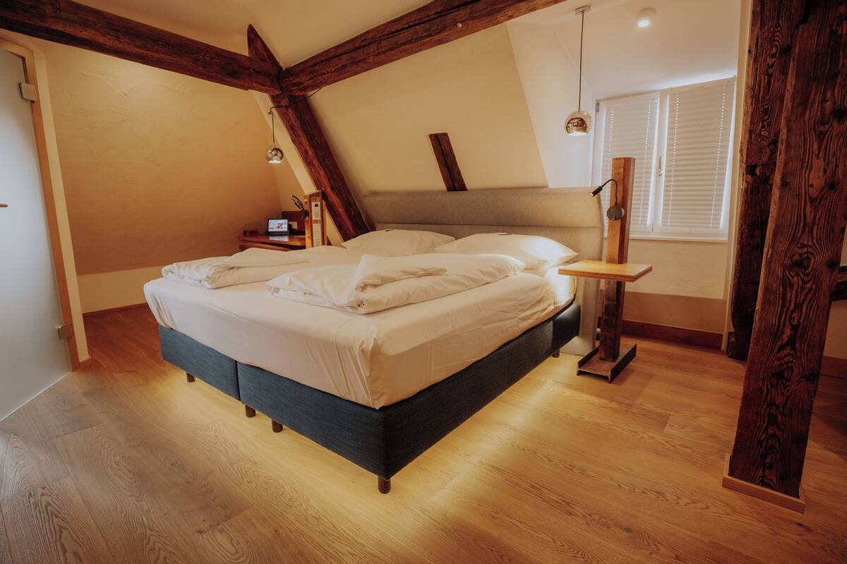 A bedroom with a bed, wooden beams, a bedside table, a lamp, and a window with blinds.