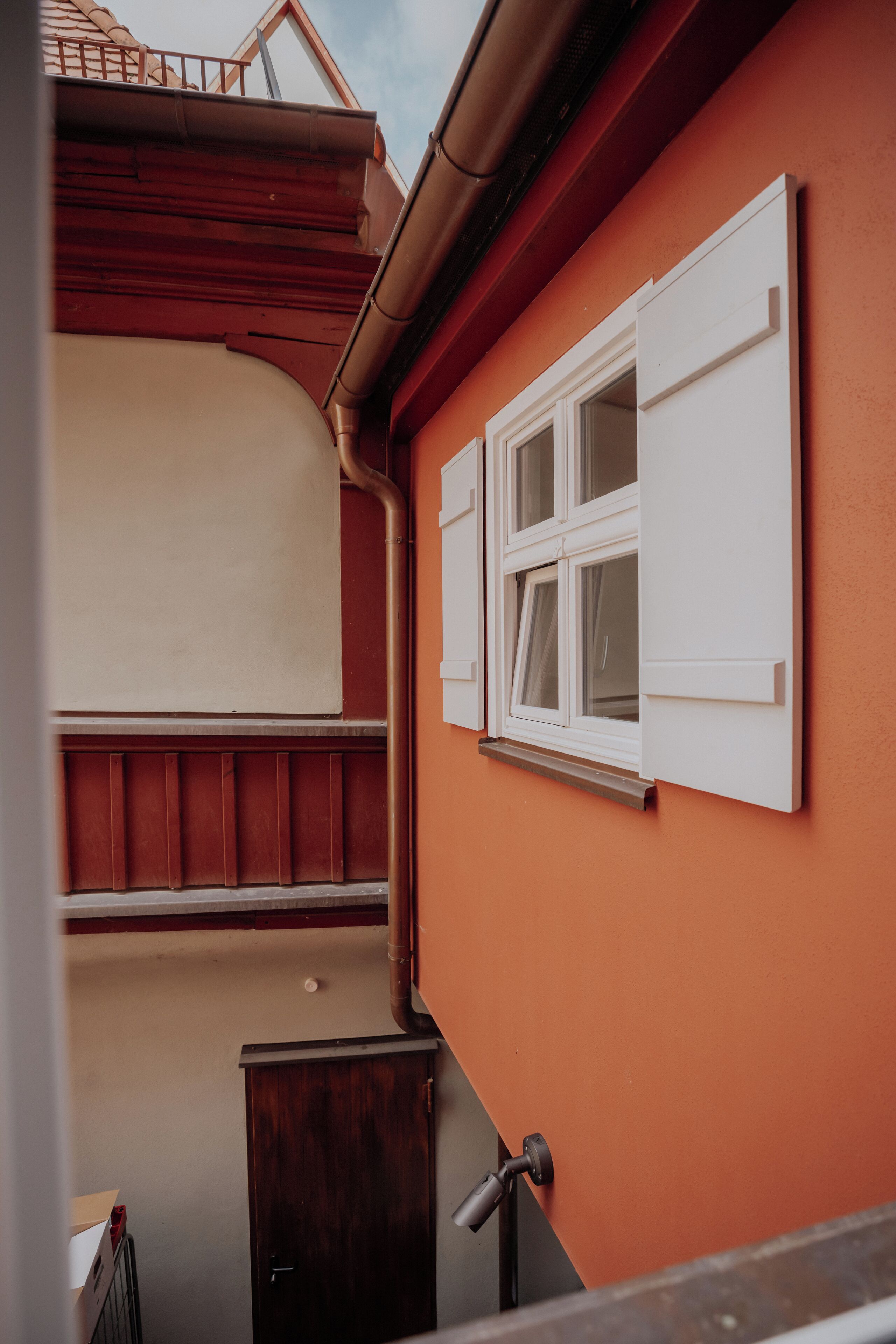 Una esquina de un edificio con pared exterior roja, una ventana con contraventanas blancas y una puerta de madera.