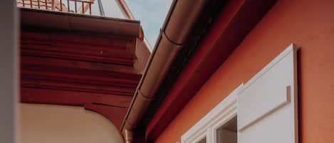 Una esquina de un edificio con pared exterior roja, una ventana con contraventanas blancas y una puerta de madera.