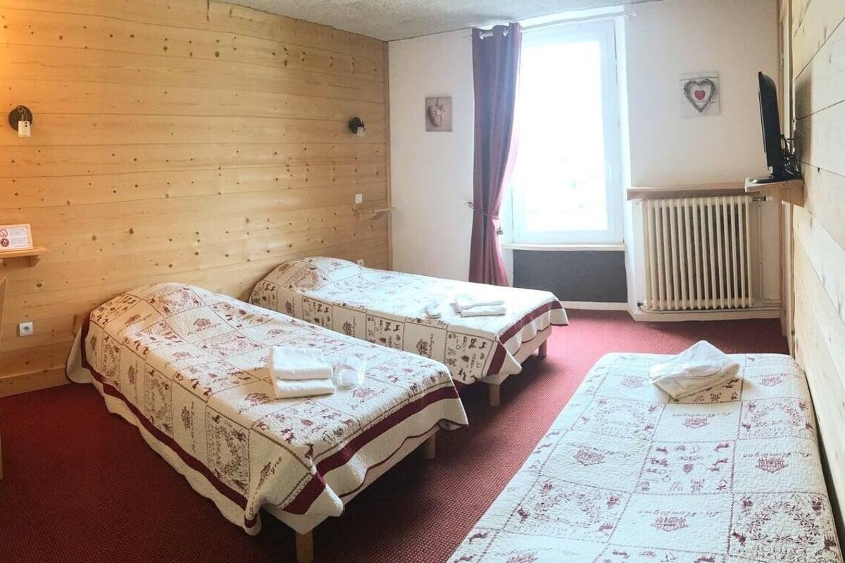 A room with two beds, a wooden wall, a radiator, a window with curtains, a small table with a telephone, and a wall-mounted TV.