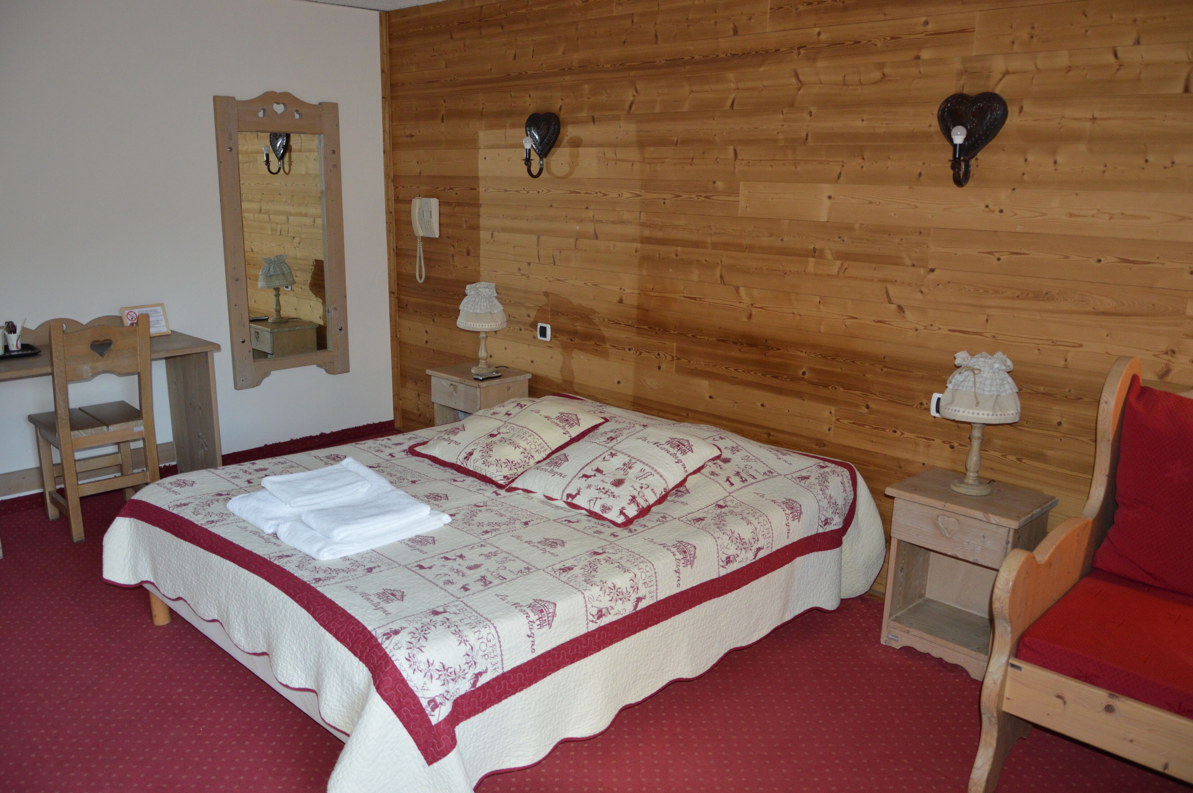 Une chambre avec un mur en bois, un lit recouvert d’une couverture rouge et blanche, une chaise en bois, une table de chevet avec une lampe et un miroir.