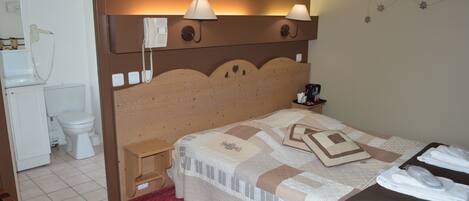 Une chambre d’hôtel avec une tête de lit en bois, un lit recouvert d’une courtepointe, une table de chevet avec une lampe, un petit tabouret en bois et une salle de bain avec des toilettes et un lavabo.