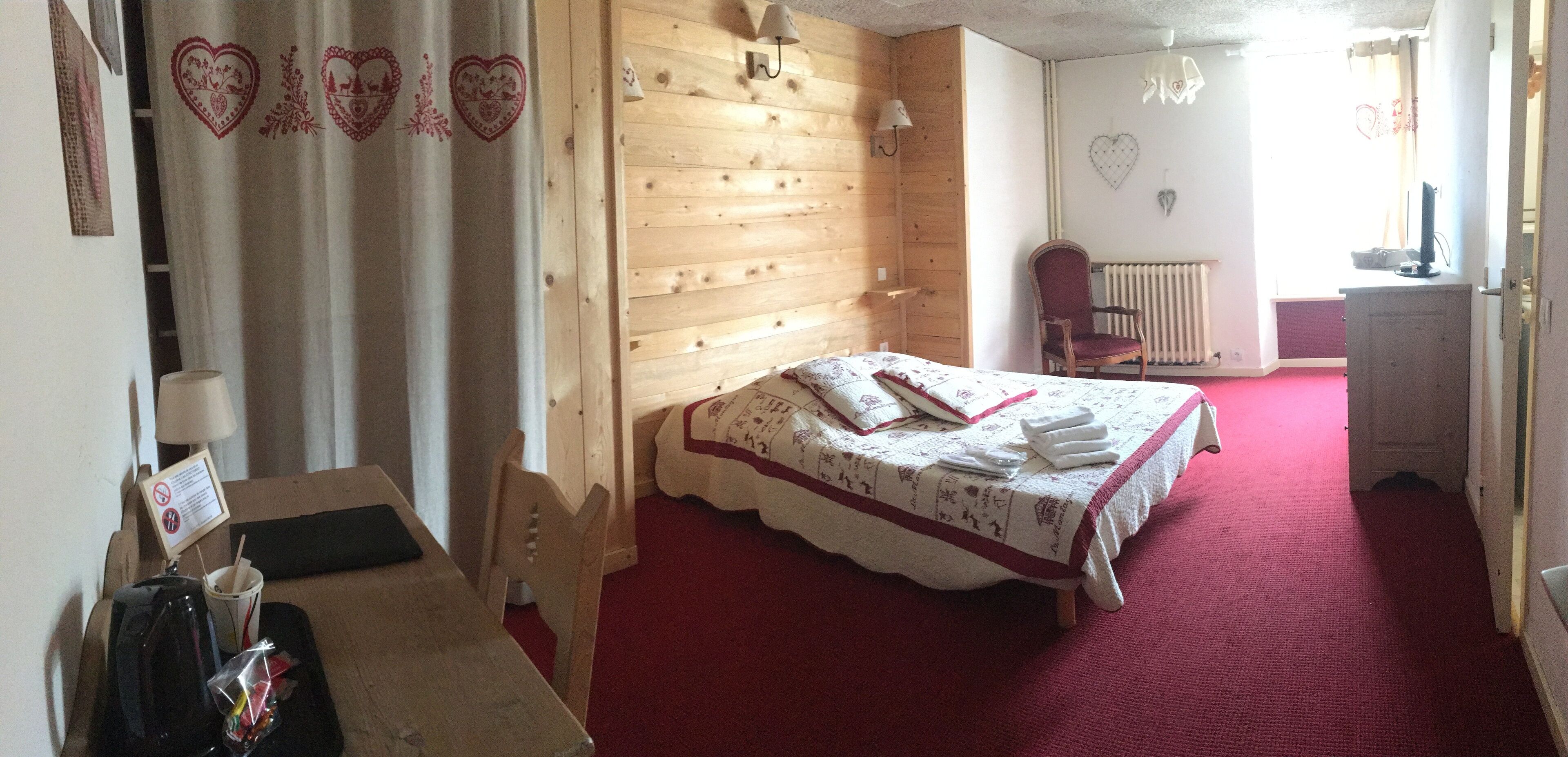Een gezellige kamer met een houten hoofdeinde, een bed met een rood-witte dekbedovertrek, een stoel en een klein tafeltje met een lamp en een kop.