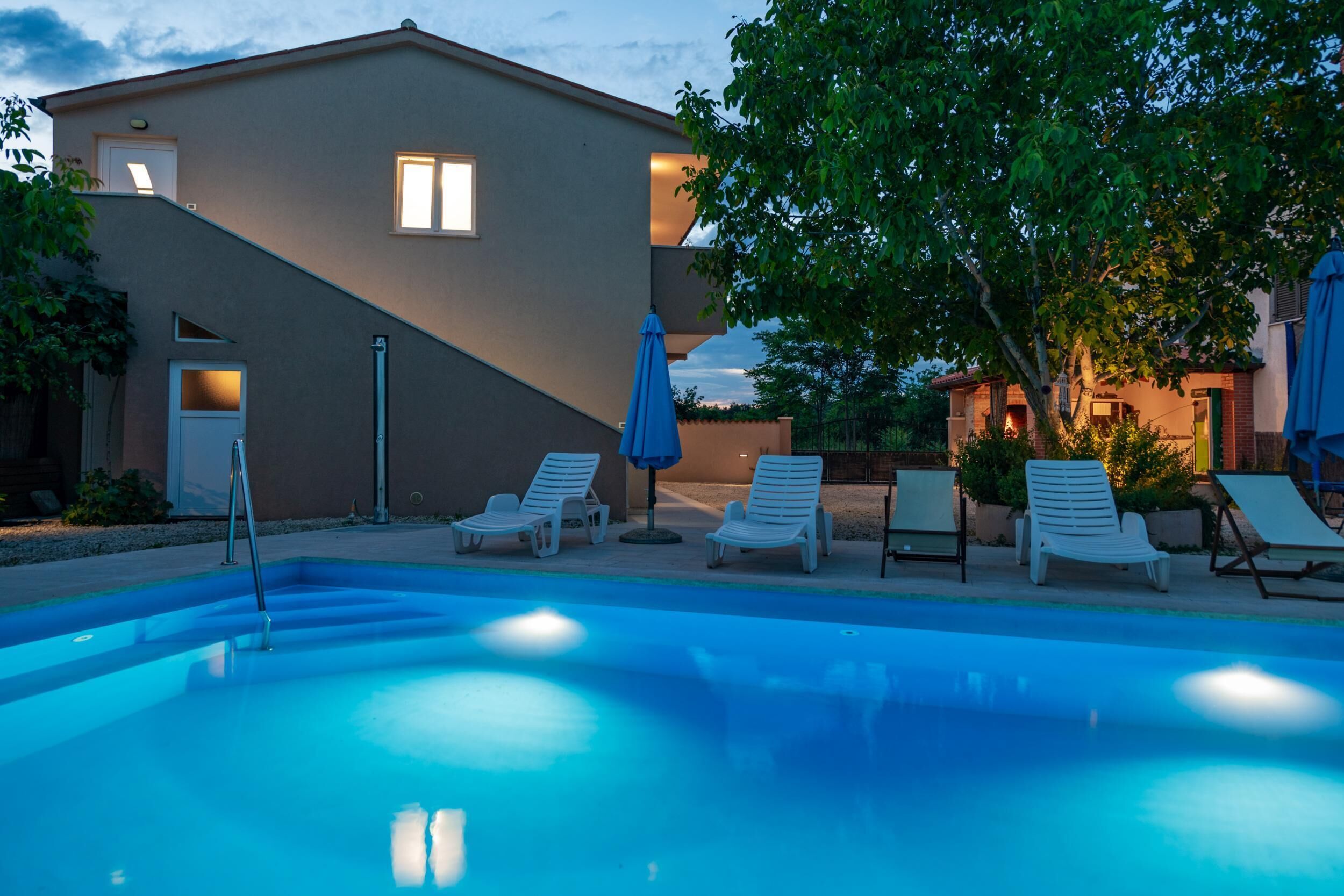 A swimming pool with lounge chairs and umbrellas, a building with a sloped roof, and a tree in the background.