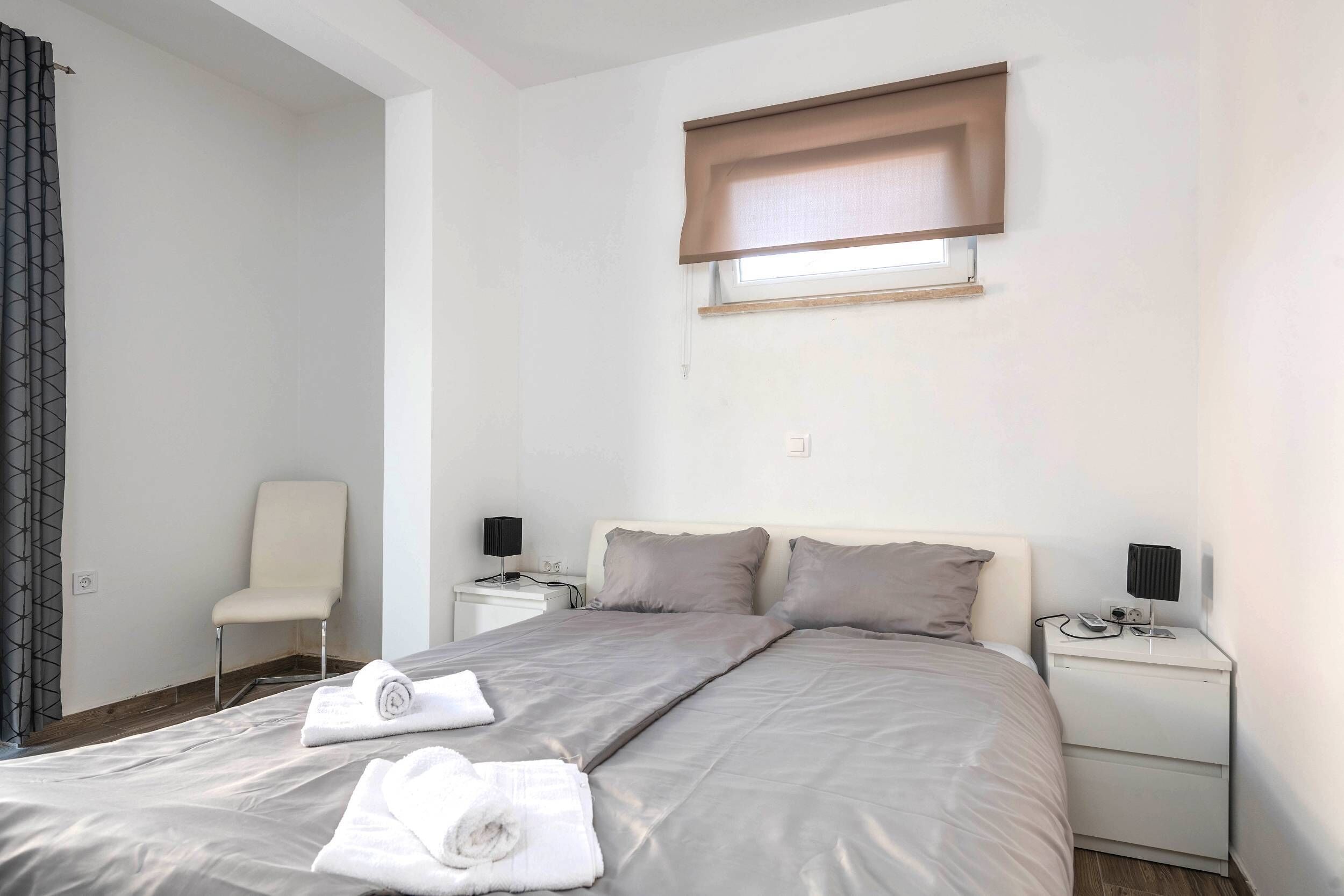 Ein Schlafzimmer mit einem Bett, zwei Nachttischen, einem Sessel, einem Fenster mit Jalousie und einem Handtuch auf dem Bett.