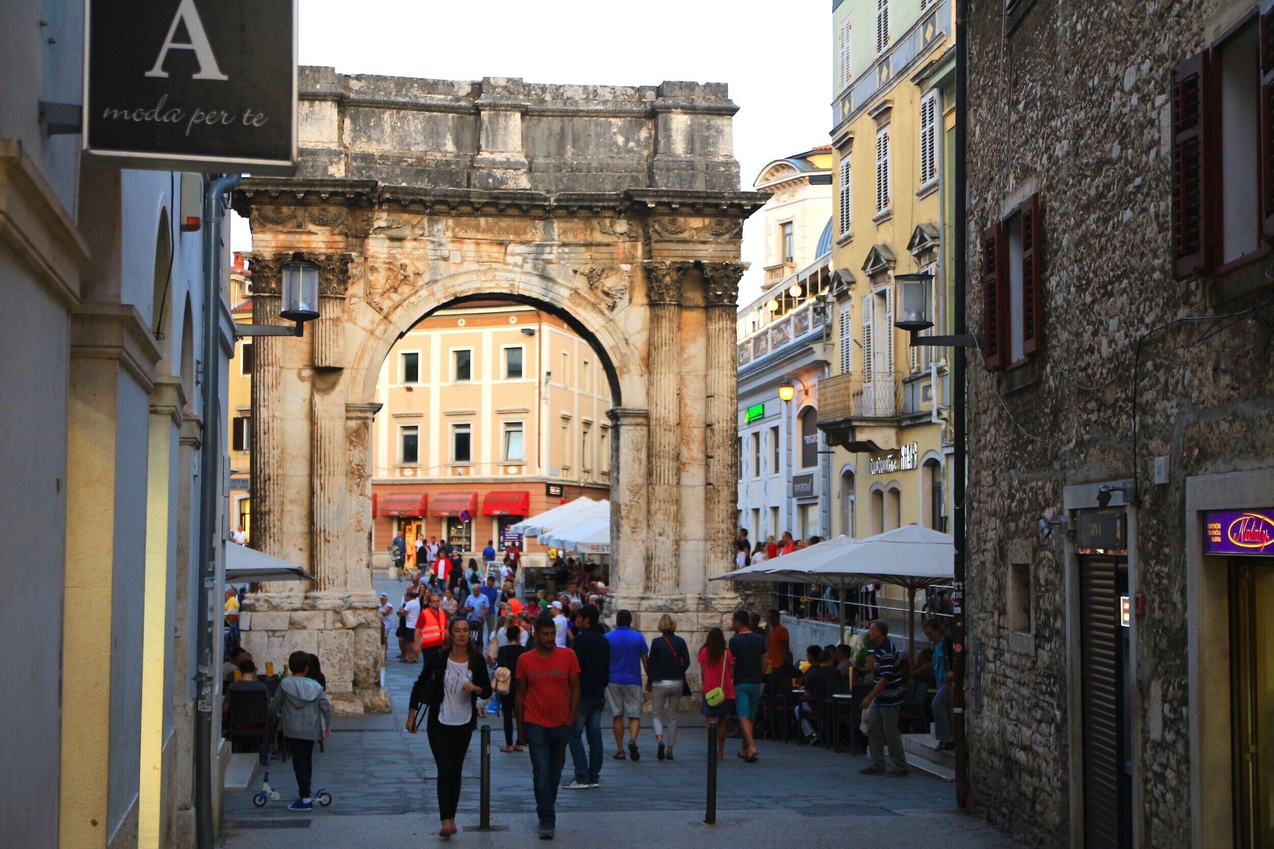 A bustling street with an ancient archway, shops, and outdoor seating areas.