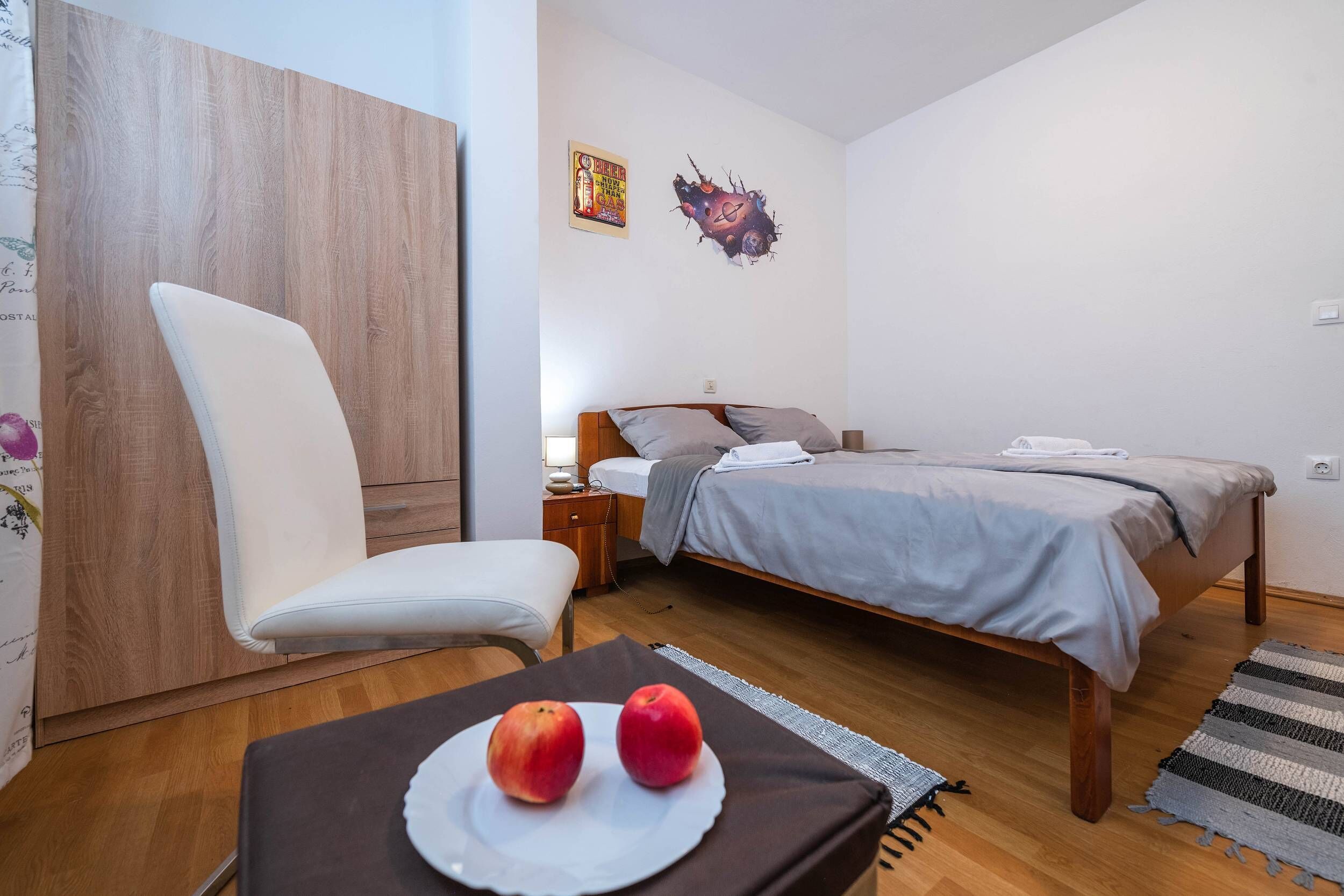 A bedroom with a bed, a chair, a small table with apples, a nightstand, and a wall decoration.