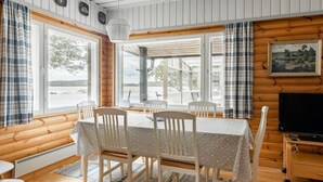 A cozy cabin with a dining area featuring a table and chairs, large windows with curtains, a painting on the wall, and a television on a wooden stand. - Wuorilahti by Interhome (Savonlinna)