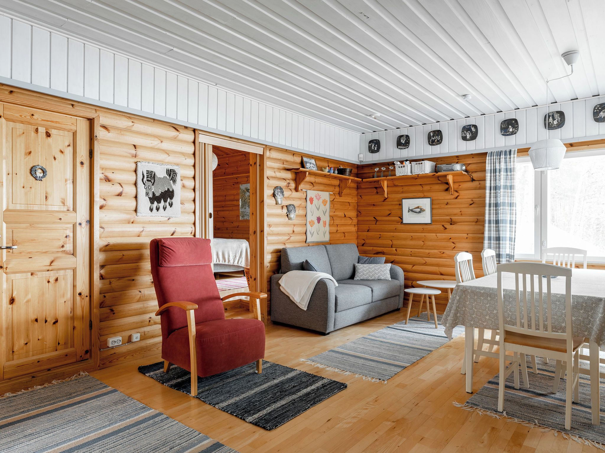 L’intérieur d’une cabane en rondins, avec une chaise rouge, un canapé gris, une table à manger en bois et un coin cuisine doté d’étagères.