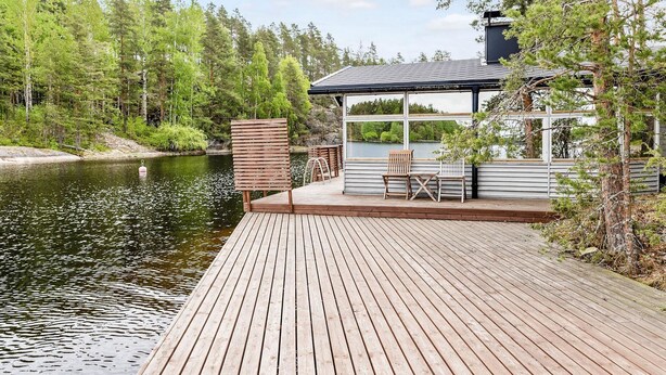 A wooden deck extending over a body of water, with a house featuring large windows and a covered patio area. - Wuorilahti by Interhome (Savonlinna)