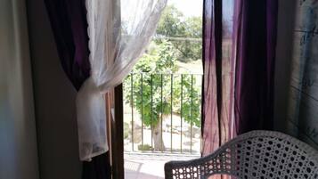 A room with a bed, a wicker chair, and a view of a balcony with a railing and trees.
