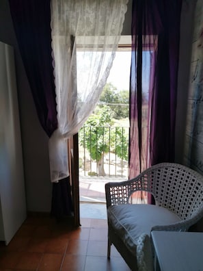 A room with a bed, a wicker chair, and a view of a balcony with a railing and trees. - Al passo di Talia (Ricadi)