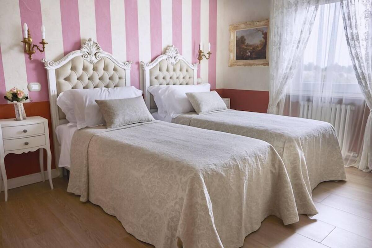 A room with two beds, each with a white headboard and matching bedding. There is a nightstand with a lamp and a vase of flowers. The walls have a pink and white striped pattern, and there is a window with sheer curtains.