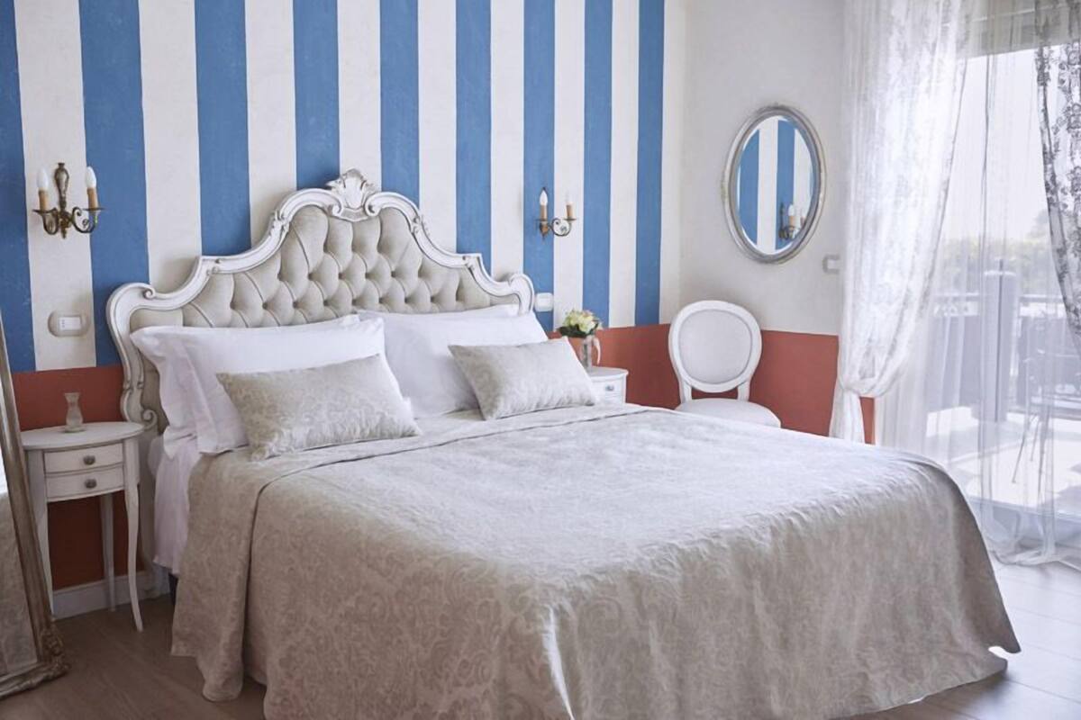 A bedroom with a striped wall, a tufted headboard, a round mirror, and a bedside table with a vase.