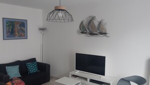 Un salon moderne avec un canapé noir, un téléviseur à écran plat, un luminaire suspendu et des œuvres d’art murales représentant des voiliers.