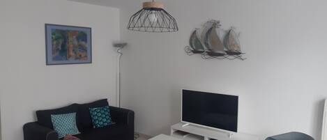 Uma sala de estar moderna com um sofá preto, uma TV de tela plana, uma luminária pendente e arte decorativa de um veleiro na parede.