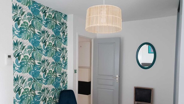 Une pièce dont le papier peint est orné de motifs de feuilles tropicales, avec une porte en bois, une chaise bleue, une petite table en bois et un luminaire suspendu.