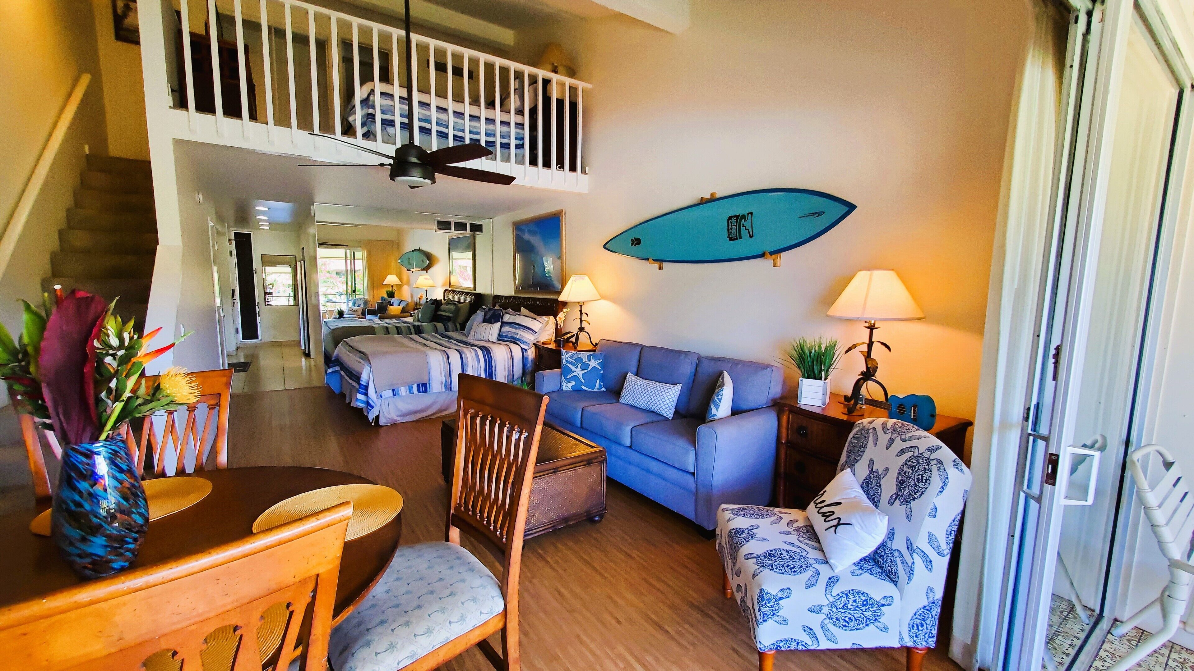 Un grand salon comprenant un canapé, des fauteuils et une table à manger. On peut y voir un ventilateur de plafond et une décoration sur le thème du surf.