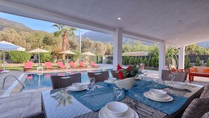 Area pranzo a bordo piscina con tavolo apparecchiato per due, circondata da poltrona a sdraio e ombrelloni, con vista su una piscina e un paesaggio montuoso.