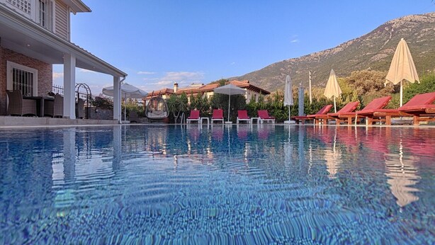 Una piscina con lettini e ombrelloni, circondata da edifici e montagne.