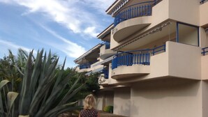 Un edificio de varios pisos con balcones, rodeado de plantas grandes.
