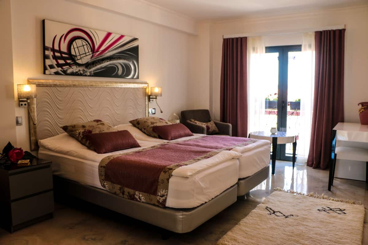 Un lit double avec une tête de lit, deux tables de chevet, une chaise, une petite table et une fenêtre avec des rideaux.