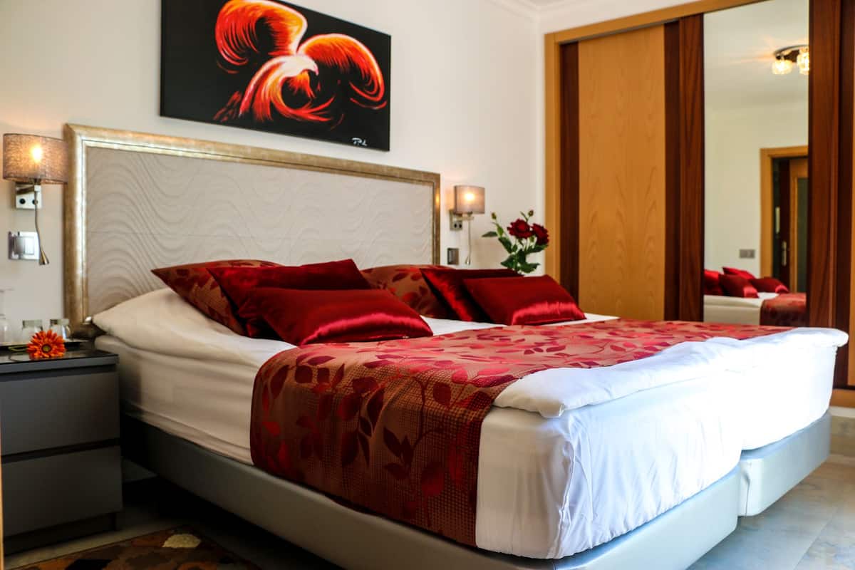 Un grand lit avec une literie rouge et blanche, une table de chevet avec une lampe, un tableau au-dessus du lit et un miroir sur le mur.