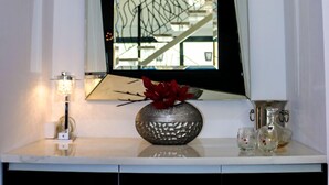 Un miroir avec un cadre géométrique, un vase rempli de fleurs rouges et une petite lampe posée sur une console blanche.