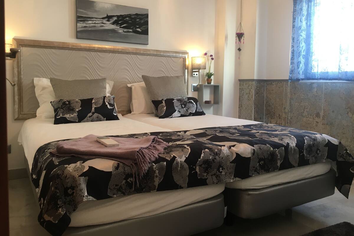 Une chambre à coucher avec un grand lit, une tête de lit, une œuvre d’art accrochée au mur, une table de chevet avec une lampe et une fenêtre avec des rideaux.