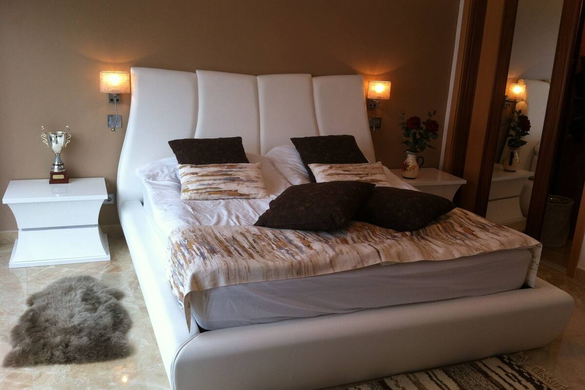Une chambre moderne avec un grand lit, une tête de lit blanche et de nombreux oreillers. On y trouve des tables de chevet avec des lampes, et une étagère où est exposé un trophée.