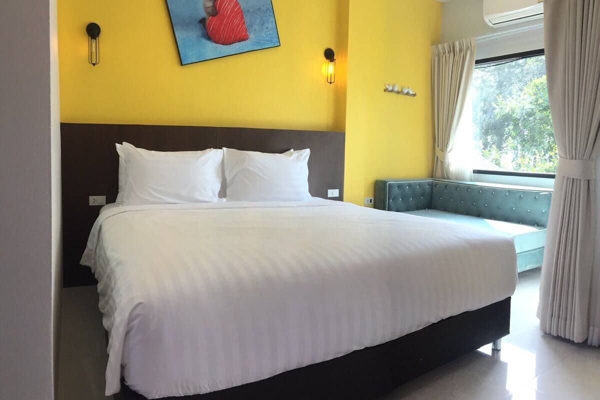 Hotelkamer met een groot bed, een hoofdeinde, een nachtkastje, een lamp, een schilderij aan de muur, een raam met gordijnen en uitzicht op bomen.