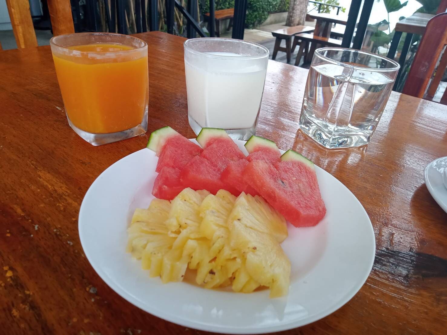 Uma mesa de madeira com uma fatia de abacaxi e melancia, dois copos de suco de laranja e um copo de leite.