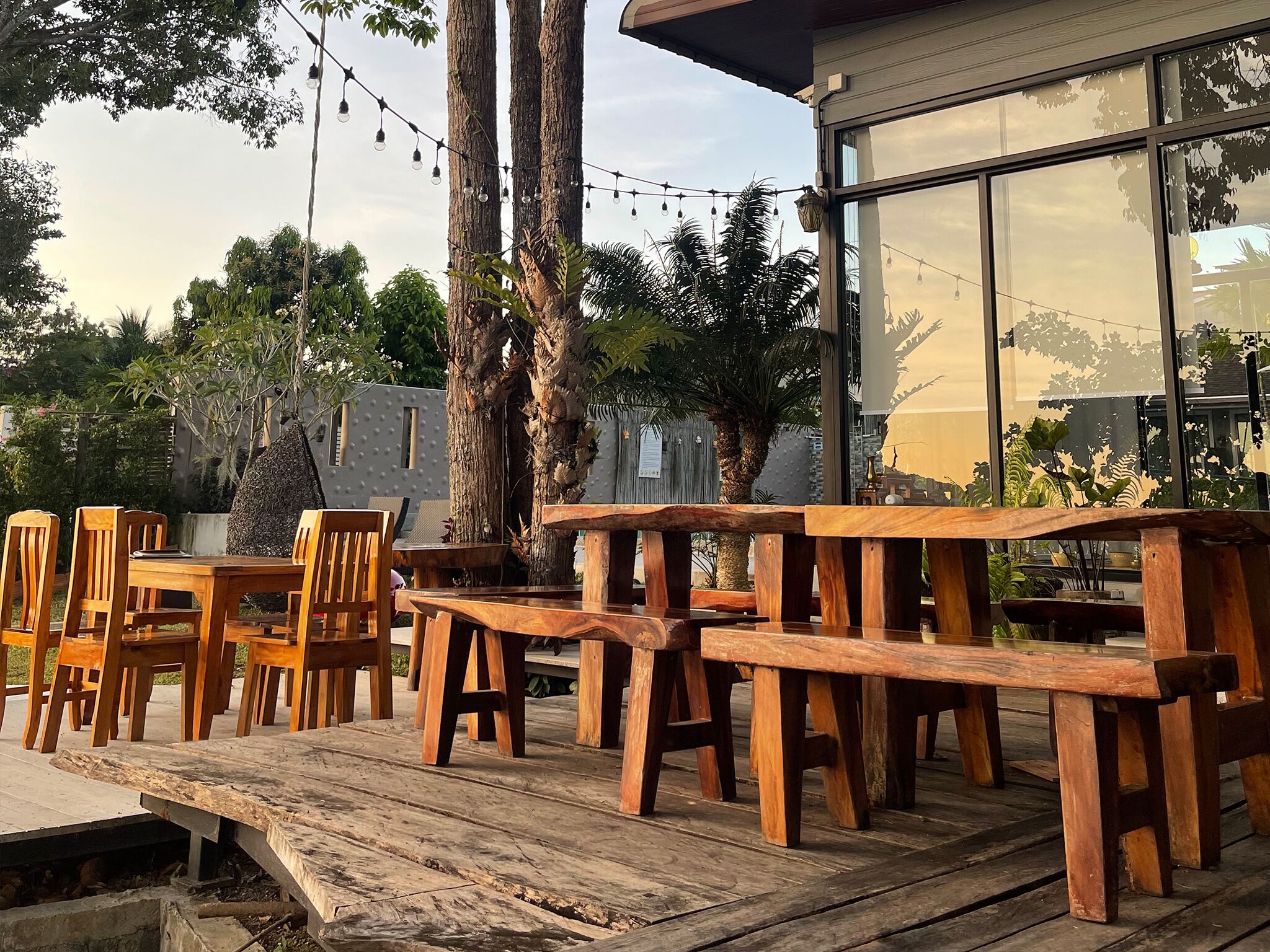 An outdoor seating area with wooden tables and chairs, surrounded by trees and a building with large windows.