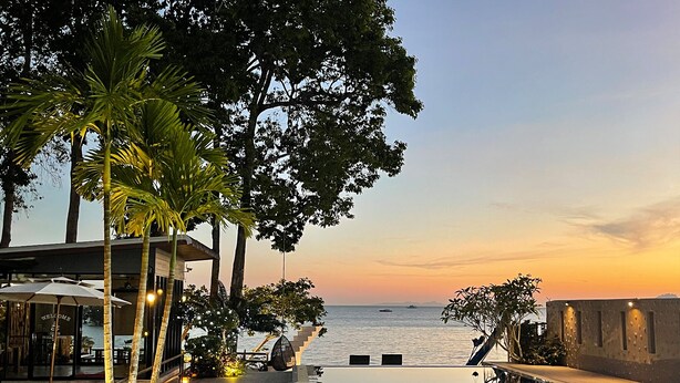 A poolside area with a curved pathway leading to a pool, surrounded by palm trees and a view of the sea at sunset. - Blue Orchid Beach Krabi (Krabi)