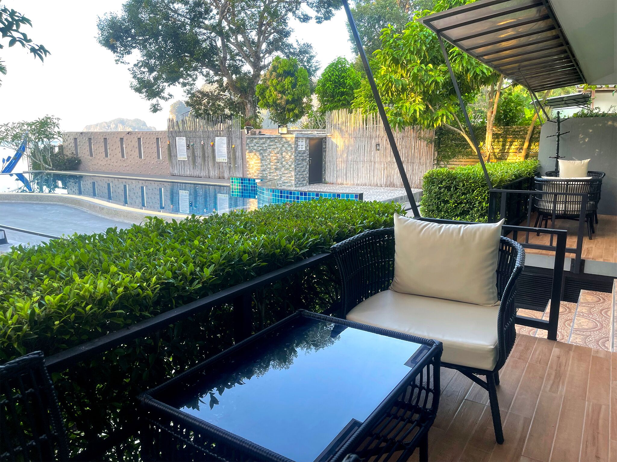 Eine Dachterrasse mit einem Glastisch und schwarzen Korbstühlen, im Hintergrund ein Schwimmbecken und Blick auf ein Gebäude mit einer gemusterten Fassade.